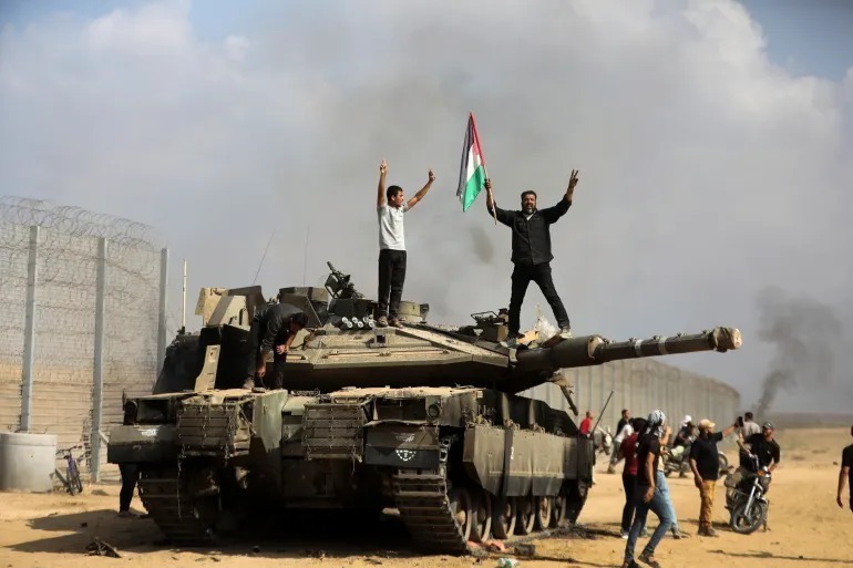 Palestinians wave their national flag and celebrate by a destroyed Israeli tank at the Gaza Strip fence east of Khan Younis on October 7, 2023. Image: AP.