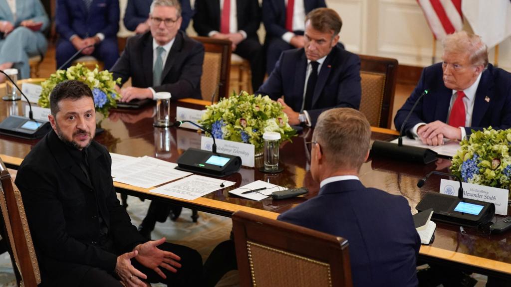 The image depicts a significant diplomatic moment at the White House where Ukrainian President Volodymyr Zelensky is sitting at the table with U.S. President Donald Trump and various European leaders. The scene shows a formal setting inside the White House, with Zelensky dressed in a black suit rather than his usual military attire, signaling a diplomatic. Image Credit:BBC World