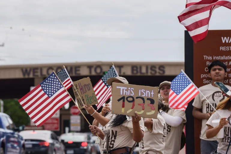 Protesters holding American flags and signs that read “This Isn’t 1933” and “#Human Not Alien” stand outside the Cassidy Gate entrance of Fort Bliss, El Paso, Texas, during a demonstration against mass deportations and a new ICE detention facility.
