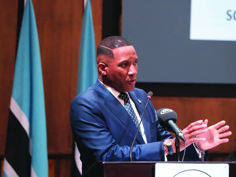 A man in a formal blue suit gestures while speaking at a podium with microphones, positioned in front of Botswana flags and a presentation screen; the scene depicts President Duma Boko during a formal announcement.