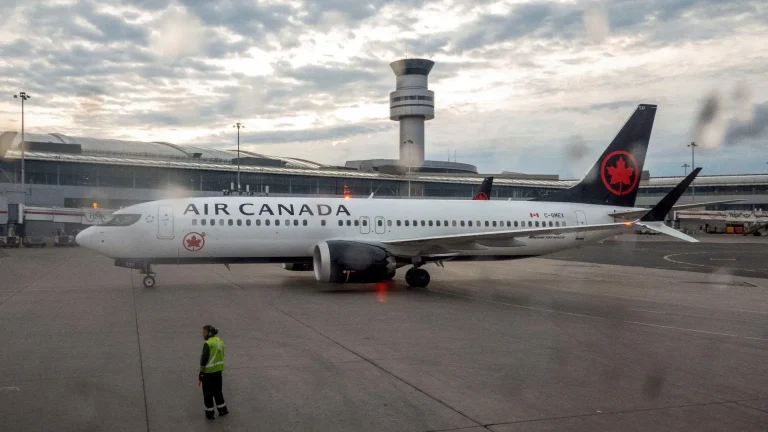 Image Credit:BBC Air Canada commercial airplane parked on airport apron near terminal and control tower, with airport ground crew member wearing a high-visibility vest standing nearby under a cloudy sky.