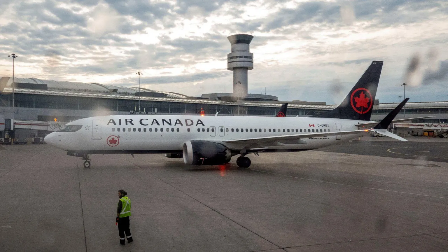 Image Credit:BBC Air Canada commercial airplane parked on airport apron near terminal and control tower, with airport ground crew member wearing a high-visibility vest standing nearby under a cloudy sky.