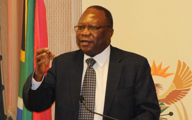 Image Credit:Photo courtesy of the World Affairs Council A formal portrait of the late Johannes Mahlangu, South African ambassador to Kenya, speaking at a podium. He is wearing a dark suit, white shirt, and patterned tie, with glasses. The background includes South African flags and an emblem.