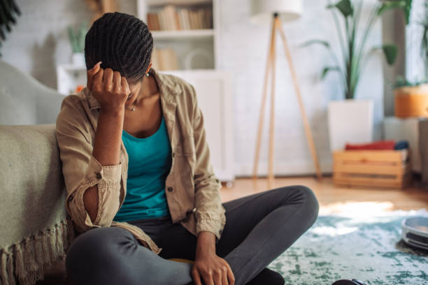Depression in Black communities is illustrated by a Black woman sitting on the floor, leaning against a couch with her head in her hand, visibly overwhelmed and emotionally distressed.