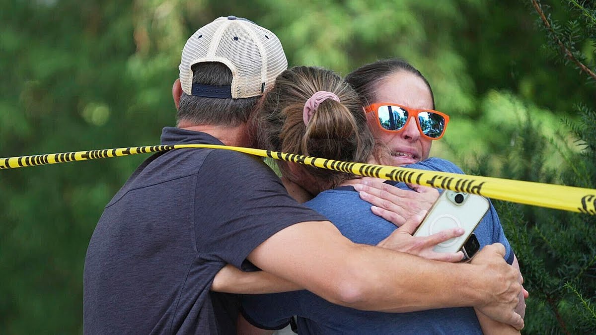 Minneapolis School Shooting: A group of people, including a woman wearing orange sunglasses, embrace each other in distress while standing behind a police cordon.