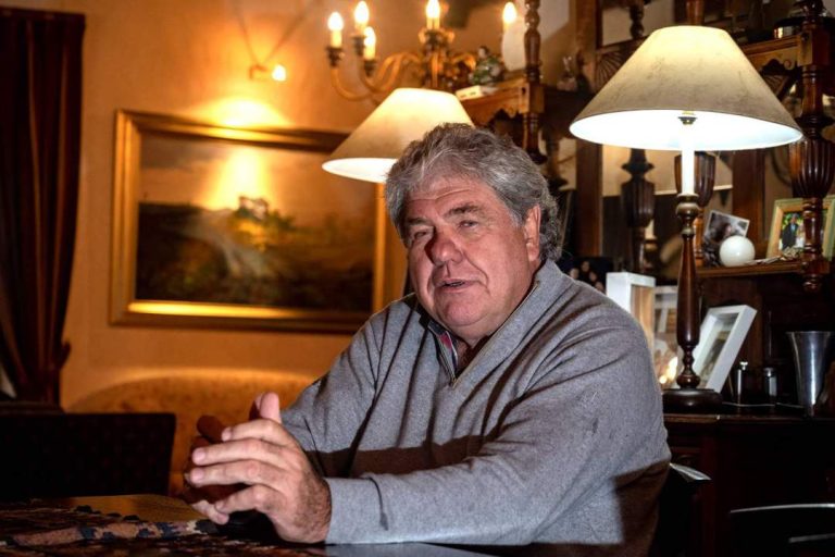 Schalk Burger seated indoors, speaking at a table, with warm lighting and a landscape painting in the background.