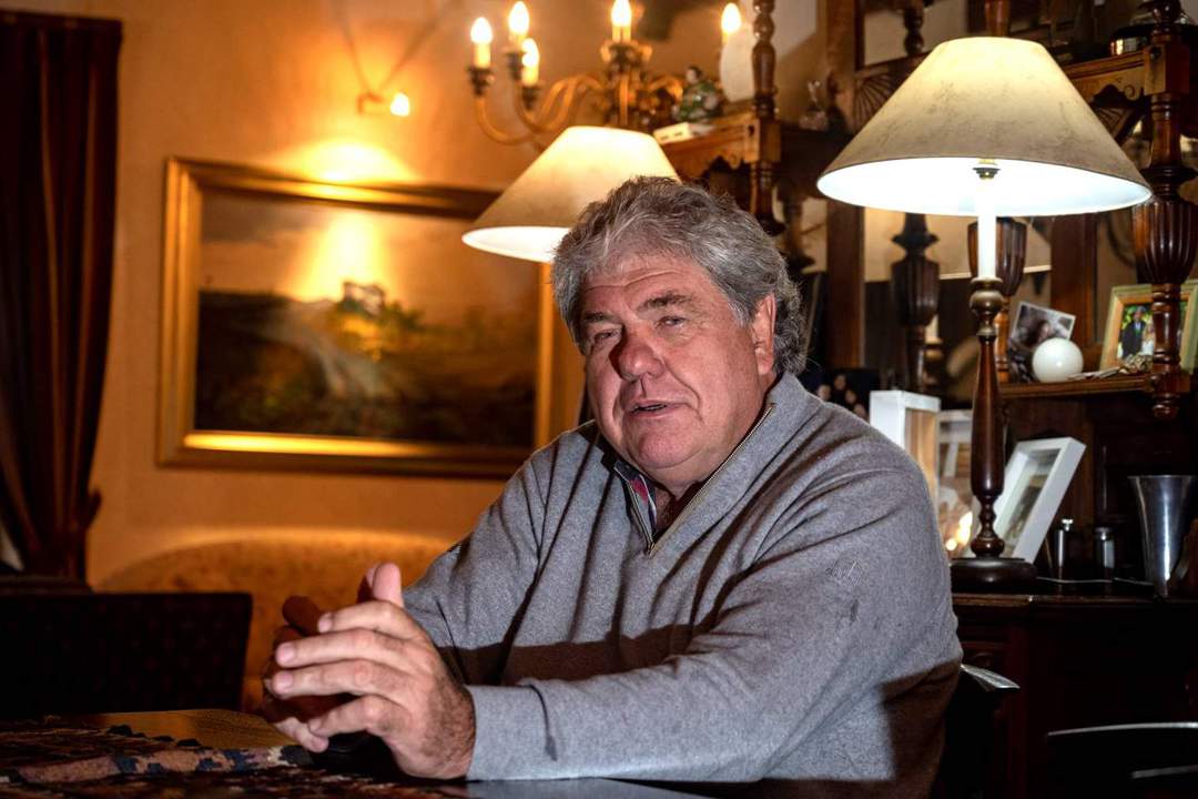 Schalk Burger seated indoors, speaking at a table, with warm lighting and a landscape painting in the background.