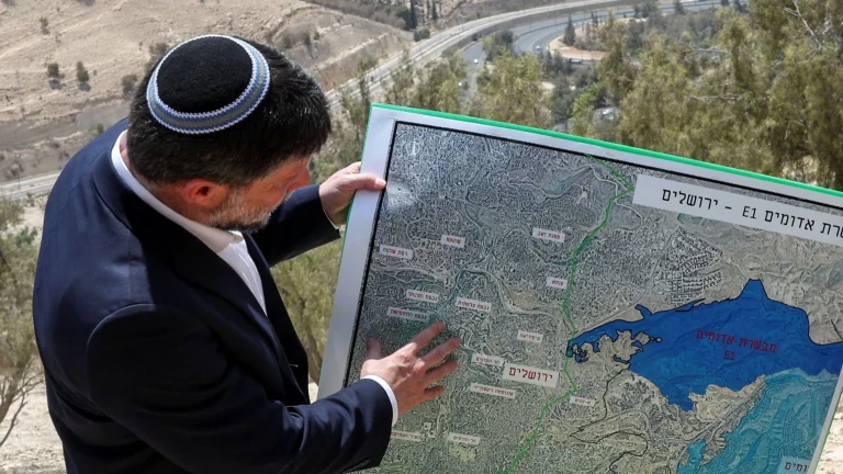 Credit Image: Reuters A man wearing a kippah and a dark suit pointing at a detailed map outdoors, showing a region with labeled areas and a large blue body of water, with trees and a road visible in the background.