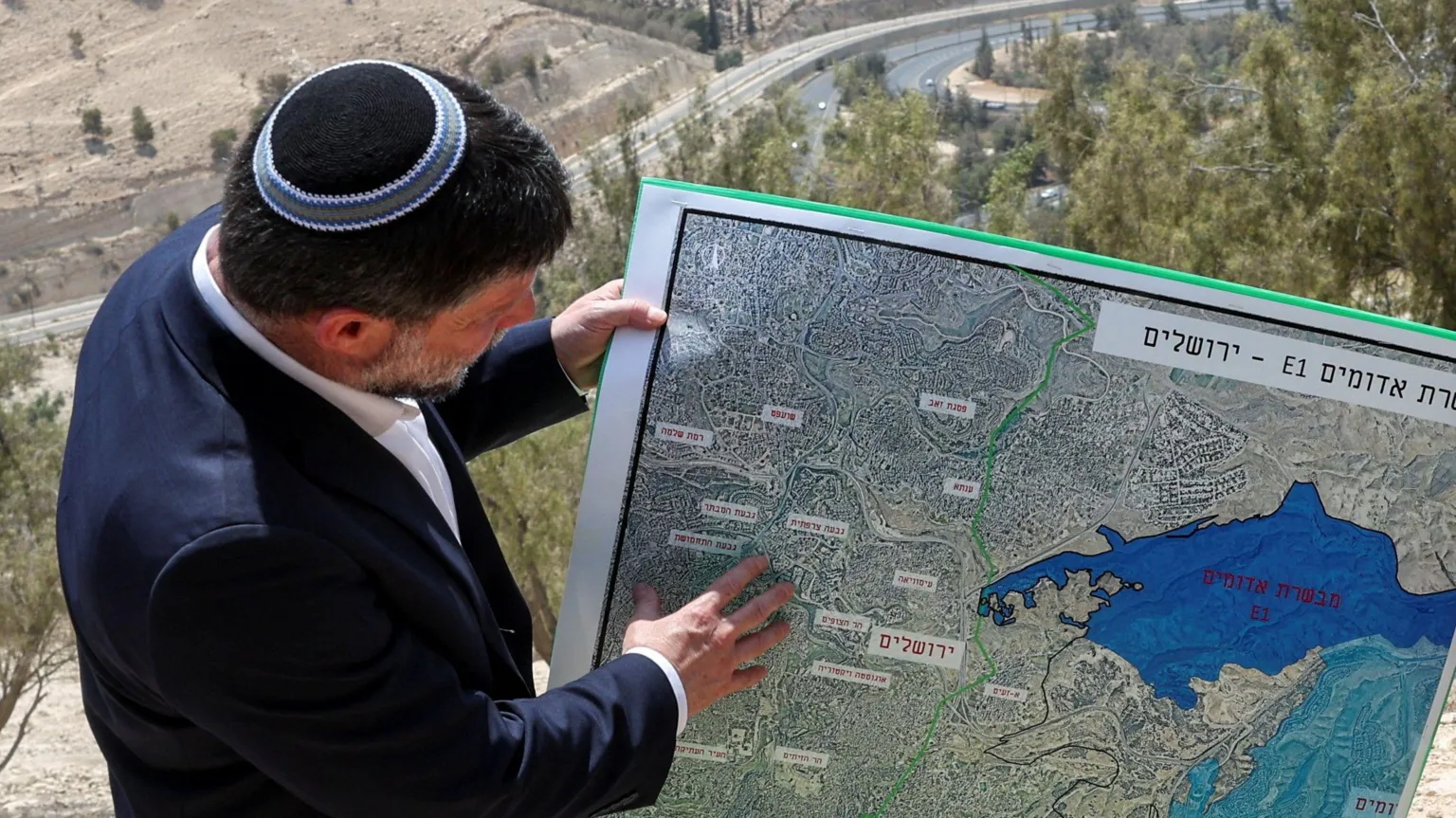 Credit Image: Reuters A man wearing a kippah and a dark suit pointing at a detailed map outdoors, showing a region with labeled areas and a large blue body of water, with trees and a road visible in the background.