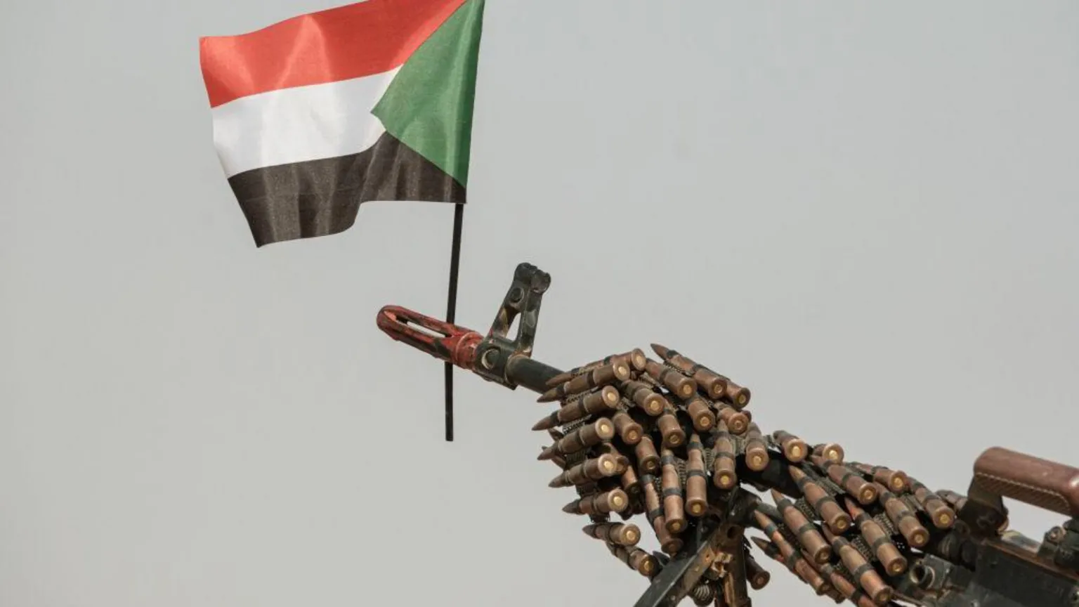 Close-up of a machine gun barrel wrapped with linked ammunition with the Sudanese flag attached to the muzzle against a plain background, symbolizing armed conflict in Sudan, particularly related to violence in Darfur.
