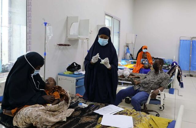 Image Credit:Reuters Diphtheria patients receive treatment inside a ward at De Martino Public Hospital in Mogadishu, Somalia. In the foreground, a woman wearing a long black dress, black covering with only her eyes visible, and a blue mask carries a child. Another woman, also dressed in a long black dress and traditional Muslim head covering, wears white gloves and holds an injection, preparing to treat patients amid the diphtheria outbreak.