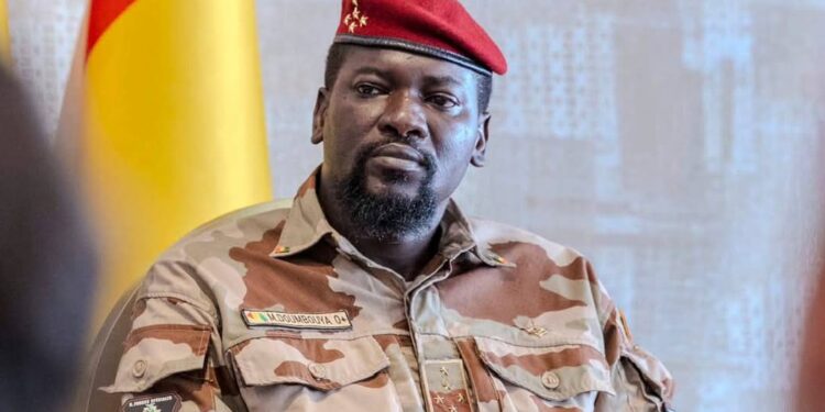 The president of Guinea in military uniform and red beret sits indoors with the Guinea flag, connected to media ban lifting.