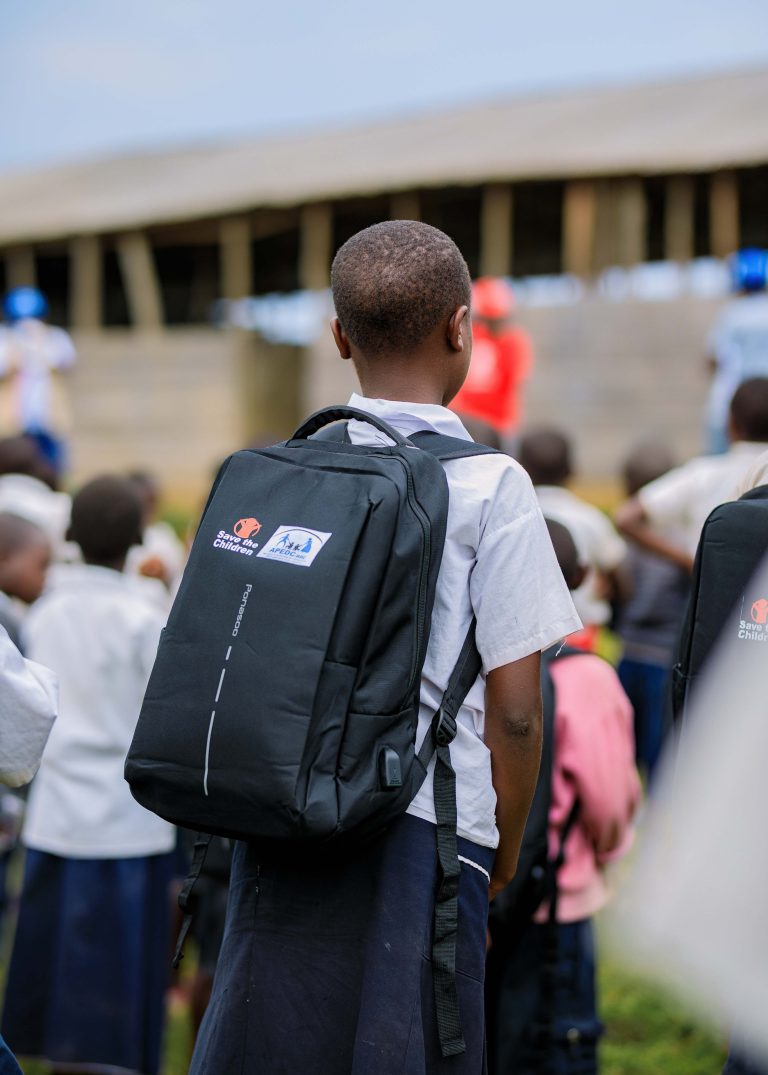 School children in eastern DRC wear blue skirts, white t-shirts, and pink jerseys, carrying blue "Save the Children" schoolbags. They study amid insecurity caused by the M23 rebel group controlling parts of the region, disrupting education and daily life.