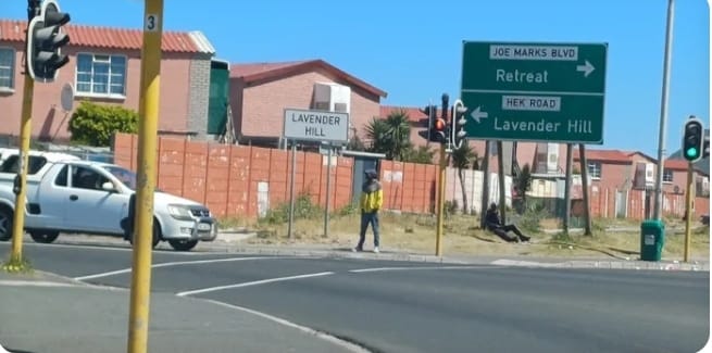 Cape Flats Chaos: A street scene in Lavender Hill, Cape Town, with road signs pointing to Retreat and Lavender Hill, and pedestrians walking past. The area is part of the Cape Flats, often in the news due to rising gang violence and community unrest.