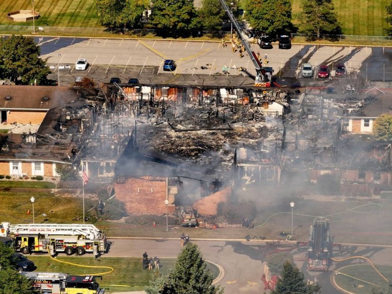 Aerial view of a burned church building in Michigan following a tragic attack; firefighters and emergency vehicles are on the scene with smoke still rising from the ruins.
