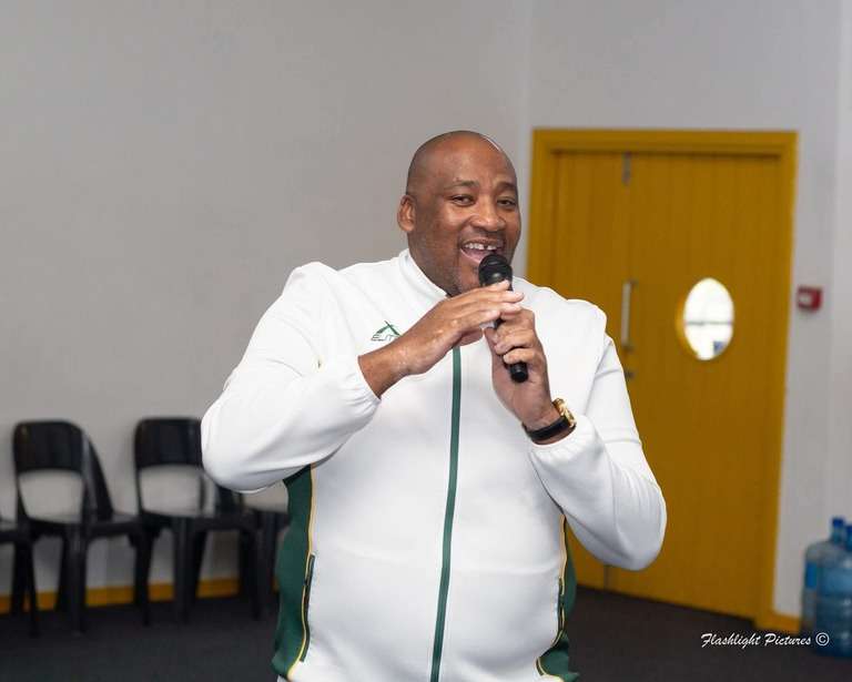 South Africa arts funding cuts: Minister Gayton McKenzie speaks at a public event, holding a microphone and smiling, wearing a white and green jacket.