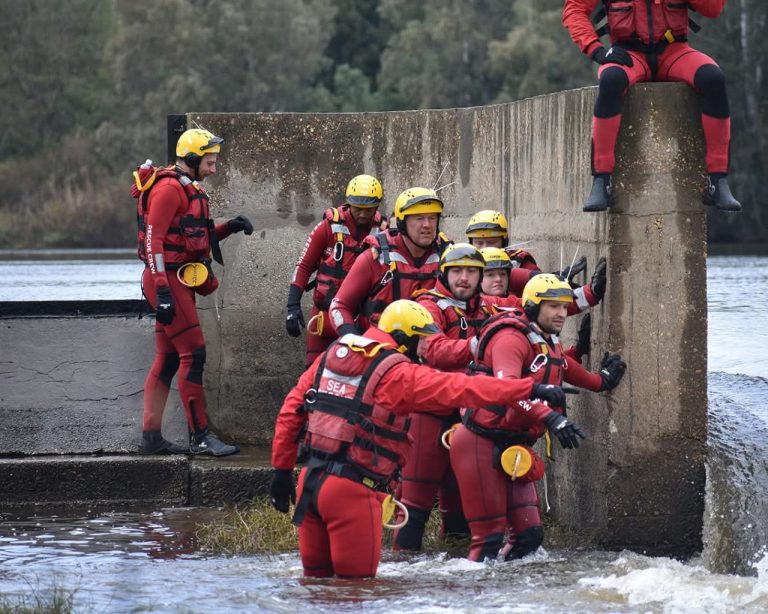 Rescue teams in red wetsuits and yellow helmets gather at a concrete structure in the water during a search operation for a missing Delft 18-year-old.