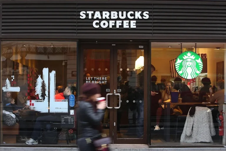 People sitting inside a busy Starbucks coffee shop, seen through the glass storefront with holiday decorations.