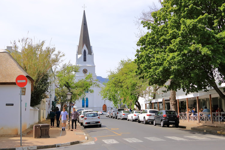 Stellenbosch Racism Probe: A quiet street in Stellenbosch, South Africa, with a prominent white church tower, lined with parked cars and pedestrians, amid the backdrop of a racial discrimination investigation involving municipal leadership.