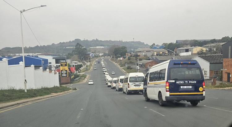 KZN Taxi Strike — A long line of minibus taxis travels down a main road in KwaZulu-Natal, with a Durban Metro Police vehicle leading the convoy, symbolizing the critical role of taxi transport in the province.