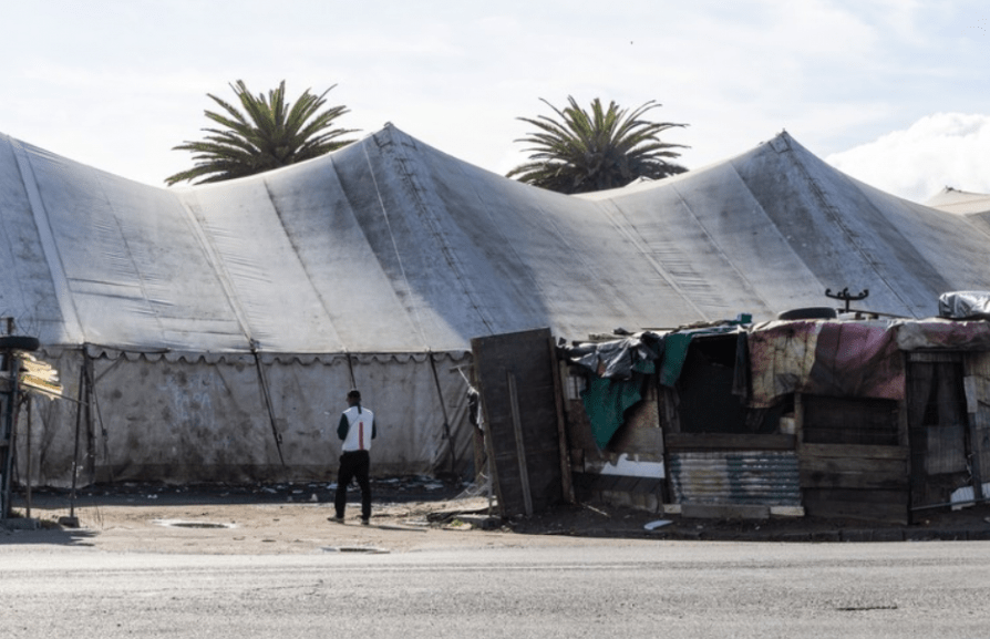 160 illegal squatters Wingfield evictions – A man stands in front of large, weathered white tents and a makeshift shack built from scrap materials, symbolizing temporary or informal housing conditions amid eviction tensions.