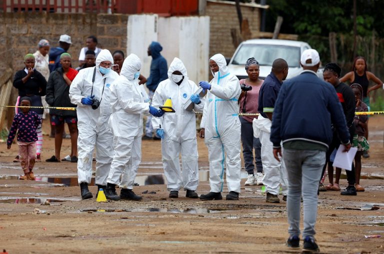 Police tape cordoning off the KwaNoxolo tavern, with community members gathered in the background, looking shocked and concerned.